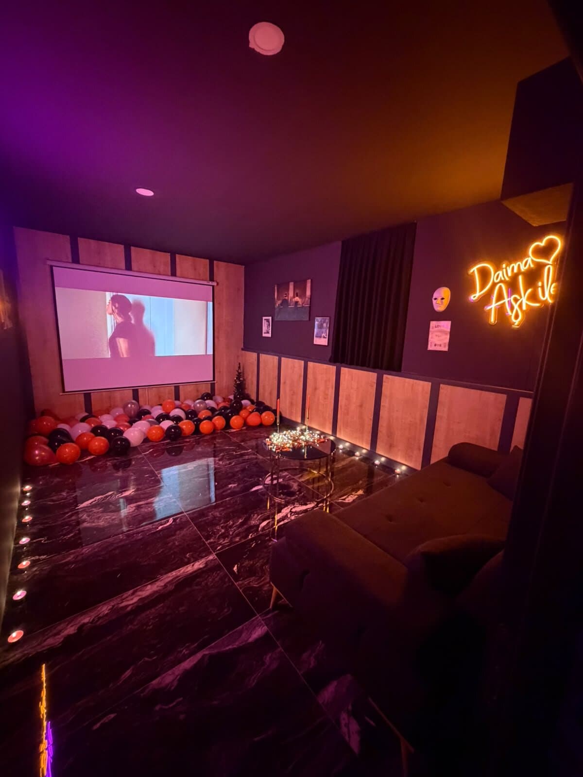 Daima Aşk ile yazan turuncu neon, perdede film, zeminde balonlar ve mum sırası, kadife koltuk